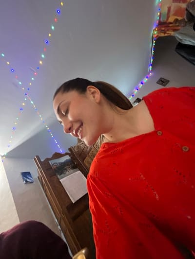 A young adult female, with brown hair styled in a ponytail, is captured in a close-up, low-angle shot within a room in Ancenis, France. She wears a red, long-sleeved top featuring eyelet patterns and three visible wooden buttons. Her head is angled downward and to the left, displaying a smile with teeth. The environment includes a light-colored ceiling and walls, illuminated by strings of multi-colored decorative lights (blue, green, red, yellow) extending across the ceiling and down a wall. In the mid-ground, a dark brown wooden screen or partition with intricate carved details is visible, upon which a calendar featuring a landscape image is affixed. A small, rectangular blue and white framed artwork hangs on a distant wall to the left. The lower-left foreground shows a partial view of a dark purple fabric object and a metallic gold-colored item.