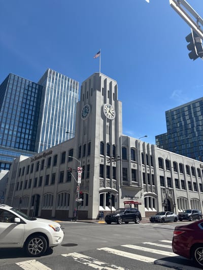 This is a low-angle, wide-shot photograph taken on a clear, sunny day in San Francisco, California, United States of America. The focal point of the image is the historic San Francisco Chronicle building, a striking Art Deco structure with a prominent clock tower. The building is made of light-colored concrete and features distinctive vertical fluting and arched windows. The American flag is flying from the top of the clock tower. To the left of the Chronicle building, two modern, tall glass skyscrapers are visible, reflecting the bright blue sky. On the right, another modern building with a dark, patterned facade is also visible.
In the foreground, a white SUV is parked or driving on the street, partially obscuring a crosswalk. Several other vehicles are also present on the street, including a silver sports car and a dark gray SUV. A red car is visible in the lower right corner of the frame. The street is paved with asphalt, and traffic signals and streetlights are present. A pedestrian can be seen in the distance, on the sidewalk near a traffic light. Traffic signs, including a "No Left Turn" sign and a sign indicating "MISSION," are also visible. The overall impression is a blend of historic and modern architecture characteristic of San Francisco.