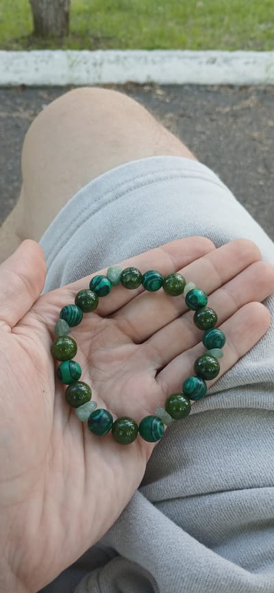 The image shows a hand holding a bracelet made of green beads. The beads appear to be malachite and possibly jade, with varying shades of green and striped patterns. The bracelet is resting on a person's palm, and the person is wearing a light grey t-shirt. The background is blurred but shows a grassy area and a dark, possibly paved, surface with a white line. The lighting suggests it is daytime, and the setting appears to be outdoors. There is no visible text in the image. The focus is on the bracelet and the hand holding it.