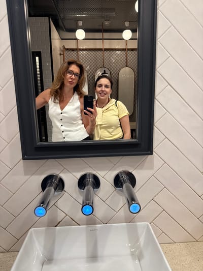 Two women are taking a selfie in a bathroom mirror in the city of Matosinhos, Portugal. The mirror is framed in dark grey and reflects the bathroom interior, showing a tiled wall and ceiling with spherical lights and a decorative metal grid. The women are holding a smartphone and looking at the camera. One woman is wearing a white top, while the other is wearing a yellow top. Next to the mirror, there's a white sink with three chrome-colored faucet handles, each featuring a blue light detail. The wall is covered in rectangular white tiles arranged in a herringbone pattern.