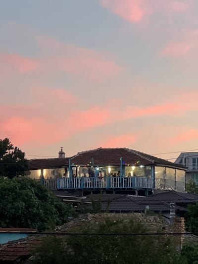 In Sinemorets, Bulgaria, during sunset, the sky displays soft pink and blue hues, with scattered clouds. A two-story building with a red-tiled roof stands prominently. The upper story features an open-air dining area with blue railings and clear plastic sheeting on the sides, illuminated by warm interior lights and colorful string lights. Several people are visible inside the dining area, seated and standing. Below this level, other buildings with dark-tiled roofs are visible, partially obscured by lush green trees. A brick chimney stands on one of the lower rooftops. To the right, a modern building with glass windows is partially visible against the sunset sky.