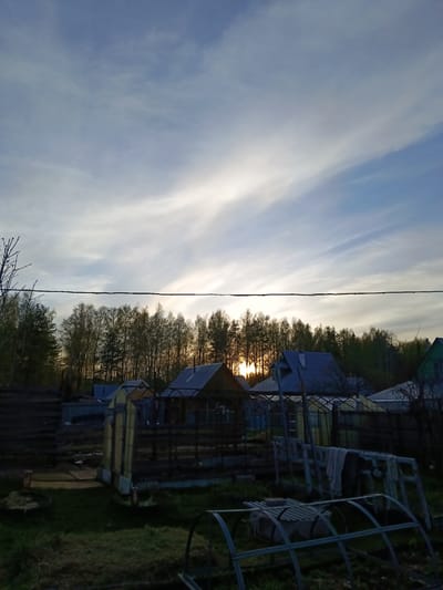 The image depicts a rural scene at dusk or dawn, possibly in the vicinity of Вичелово, Russia. The sky is a mix of blues and whites with streaky clouds, and the sun is positioned on the horizon, partially obscured by a line of trees. The silhouettes of several houses are visible in the middle ground. In the foreground, there is overgrown grass and various gardening structures, including a greenhouse frame, a possible ladder or clothes rack with hanging fabric, and other indistinct objects suggesting a backyard or garden area. A power line stretches across the upper portion of the image. The scene is generally still, and there is no visible human activity.