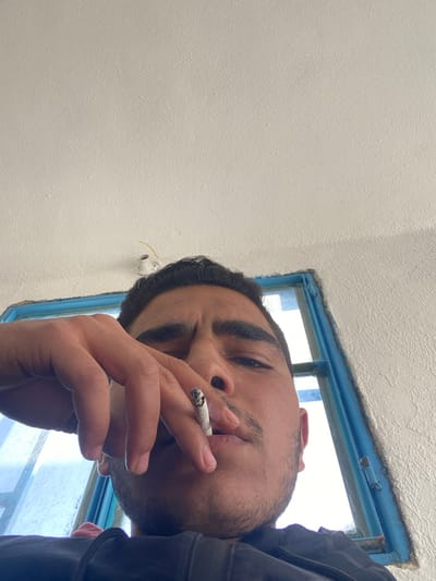 A low-angle photograph taken in Dahiyat Harasta, Syria, captures a young adult male looking directly at the camera. The male subject has dark hair, dark eyes, and light facial hair. He is wearing dark apparel. His right hand holds a lit cigarette, with visible ash, between his index and middle fingers, positioned near his mouth. The background consists primarily of a white, textured wall or ceiling. A window with a distinct blue frame is visible behind and to the right of the subject, revealing a bright, indistinct exterior. Above and to the left of the subject's head, a small, white, cup-shaped electrical fixture with yellow wiring is attached to the wall/ceiling surface.