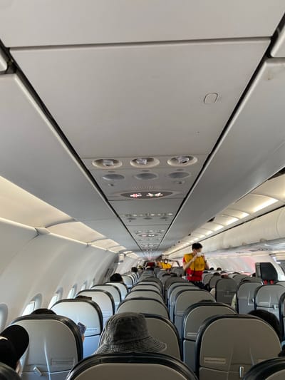 The image shows the interior of an airplane cabin. The perspective is from an aisle seat, looking forward and slightly upwards towards the overhead compartments and rows of seats. The cabin is brightly lit with overhead fluorescent lights.
Several people are visible, mostly seated. One person in the foreground is wearing a dark cap and is seen from behind. Further down the aisle, other passengers are seated, some looking at their phones. A person in a yellow shirt and red life vest is standing in the aisle, possibly attending to passengers or preparing for something.
The overhead panels feature various controls, including air vents and reading lights. The seats are typical airline passenger seats, upholstered in grey fabric with black trim. Some seats have small informational signs attached to them.
The image does not provide clear cues for the time of day or weather. However, the overall scene suggests a routine flight. The presence of a life vest on one passenger might indicate a specific type of flight or a safety drill. The location context provided is Nong Prue Subdistrict, Thailand, which may imply the flight is within or departing from Thailand. No specific emotions are discernible from the visible subjects. The visible text on the seat is a safety instruction, reading "Fasten Seat Belt While Seated".