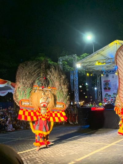 The image shows a nighttime performance in the city of Kecamatan Batu, Indonesia, specifically at the coordinates -7.867 latitude and 112.514 longitude. The image is of a street performance featuring a costumed dancer with a large, intricately-designed headdress and mask. The headdress is adorned with feathers, resembling a peacock, and there is a sign on the mask that reads "PONO ROGO". The dancer is wearing a red and yellow checkered garment with black accents and is dancing barefoot on a paved street, illuminated by a single streetlight overhead. The surrounding environment is dimly lit, and a crowd of spectators can be seen in the background, gathered in front of a large stage. The stage is covered with a white tent-like structure and is equipped with lighting and sound equipment. There is a large banner behind the stage with an image of a man and a smaller, smaller banner featuring a group of people. The dancer is performing in front of the stage. The background is filled with trees and a large, white tent-like structure.