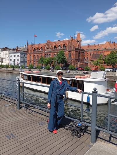 A full shot captures a person standing on a wooden walkway next to a metal railing, with a river and cityscape visible in the background. The person is wearing a denim jacket and pants, a red shirt, a white cap, sunglasses, and red shoes. They are leaning on the railing. Behind the person is a white boat with windows. In the background is a red brick building with a red flag on top, other buildings, trees, and cars. The sky is blue with scattered clouds. The location is likely in Bydgoszcz, Poland, given the architectural style and riverfront setting. A coiled black cable and blue electric plug are on the walkway near the railing.
