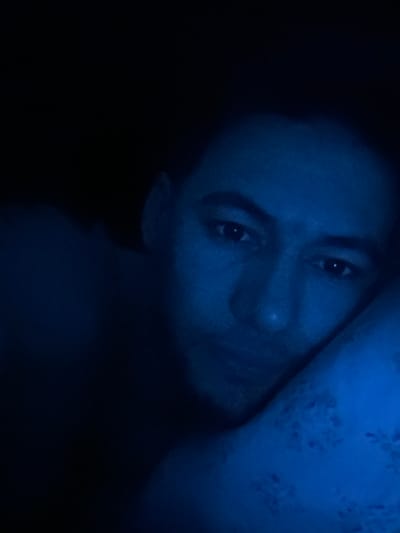 A close-up, low-light portrait of a person with short, dark hair. The image is dominated by a deep blue hue, casting a cool tone across the subject's face, emphasizing the eyes and nose. To the side, a patterned fabric in a lighter shade of blue is visible. The person appears to be lying down. A large portion of the lower half of the frame is dark and undefined.