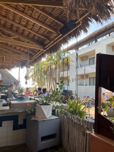 The image shows an outdoor restaurant in Brisas de Zicatela, Mexico. A thatched roof structure covers the restaurant area. A group of people are seated at tables, appearing to be dining. Outside the immediate restaurant area is a view of a building and a street where people are walking. A vehicle is also visible. The environment includes palm trees and other vegetation. The scene is a mix of restaurant activity and the surrounding urban beach environment.