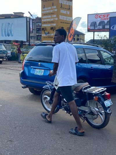 In Ogbunike, Nigeria, a man in a white t-shirt and dark shorts walks beside a motorcycle. He is wearing dark Crocs. The motorcycle is dark blue with a silver rack on the back. A blue Peugeot station wagon is parked behind the motorcycle. In the background, there are billboards advertising tires, one for "GODDARD TYRE ROADTRUSTAR TYRE" and another for "CST TIRES". A tall yellow building with the same tire advertisements is also visible. There is a street with pavement and some scattered debris. The sky is overcast with some clouds. A smaller blue truck is partially visible to the left.