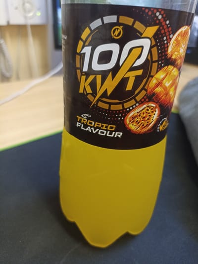 An orange-yellow liquid fills the lower portion of a clear plastic bottle, extending to its base. The bottle is labeled with a black sticker featuring a large, stylized "100 KWT" in white and gold. Images of passion fruit slices are visible on the sticker, along with text that reads "TROPIC FLAVOUR." The bottle sits on a dark surface, possibly a desk or mousepad, with a green line visible to its left. In the background, a blurred, out-of-focus office environment is suggested by a white cord and part of a piece of equipment with a green light. The scene is observed from a slightly elevated, close-up perspective. The image does not contain any elements that indicate a specific location in Shakhunya, Russia; it appears to be a generic indoor setting.