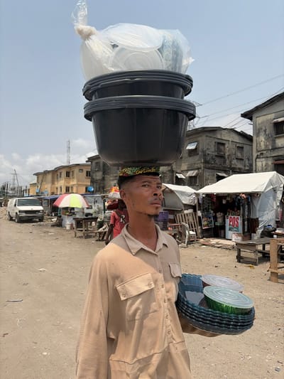 An adult male stands outdoors in Jakande, Nigeria, balancing a heavy load on his head and carrying additional items in his left hand. The man wears a light brown, long-sleeved button-down shirt with chest pockets and a patterned head-ring. On his head, stacked directly above the head-ring, are two black plastic basins, topped with a clear plastic bag filled with transparent plastic containers and lids. In his left hand, he holds a stack of multiple transparent, possibly blue or green, plastic plates/bowls with wavy edges, along with two clear lids. His gaze is directed slightly to his right, with a neutral expression.
The scene depicts an unpaved, dusty street or market area under a bright, hazy sky. In the mid-ground to the left, a white van is parked near a small market stall with a red and white striped umbrella. Further back, multi-story buildings with tan facades are visible. To the right, another market stall is covered by a white tarp, displaying goods and featuring a "POS" sign. Beyond this stall, weathered concrete two-story buildings line the street. Power lines and utility poles are visible in the background. Scattered debris is present on the ground.