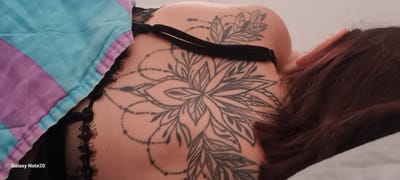 A close-up, overhead shot shows a woman's upper back and shoulder, adorned with a large, intricate black tattoo. The tattoo resembles a mandala or lotus flower, with delicate lines and shading. A black bra strap is visible across her shoulder, and a portion of her dark hair is also in the frame. The woman is lying on a quilted blanket with sections of light blue and purple. The image is watermarked "Galaxy Note20". The scene is indoors, with a neutral background that suggests a bedroom or similar private space. There is no visible indication of the city of Holon, Israel.