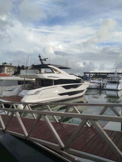 The image depicts a marina scene in Ko Kaeo, Thailand. The foreground is dominated by a wooden walkway with a metal railing. Several large yachts are moored in the water beyond the walkway. The largest yacht, in the center, is white with black trim and appears to be a luxury vessel. Other yachts of varying sizes are visible, including one with a tan canopy. In the background, buildings resembling hotels or condominiums line the shore, and a structure with what appears to be boat storage or a maintenance area is visible further back. The sky is partly cloudy with a mix of sunlight and overcast conditions. There are no people visible in the image.