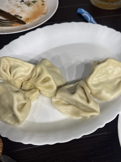 A close-up, overhead shot shows a white, ruffled plate holding five khinkali, a Georgian dumpling dish, on a dark, wooden surface. The khinkali are light beige and appear to be freshly prepared, with the dough gathered at the top. The plate has a slight sheen and is partially visible, with its scalloped edge extending towards the viewer. In the upper left corner of the image, another plate is visible, holding remnants of food, including a fork and what appears to be a reddish sauce and some green herbs. To the right of the khinkali, a portion of a glass container with a yellow liquid is visible. A blue and white patterned object, possibly a utensil or a wrapper, is also present near the top right of the frame. The lighting is dim, creating soft shadows. This scene likely depicts a meal at a restaurant in Lagodekhi, Georgia, given the presence of khinkali, a popular Georgian dish.