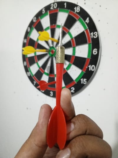 The scene depicts an indoor setting in Al Haouafate, Morocco, featuring a dartboard and darts. A standard dartboard, black, white, green, and red in color with numerical segments (e.g., 20, 18, 13, 10, 15, 2, 3, 16), is mounted on a plain white wall. Three darts are already embedded in the board: two with yellow fletching are clustered near the bullseye, one in the inner green circle and the other in the adjacent '1' segment. A third dart with red fletching is lodged in the '7' segment. In the foreground, a human hand, consistent with an adult's, grips a fourth dart, also with red fletching, its metallic point directed upwards towards the board. The dart's brass-colored knurled barrel is visible, held between the thumb and forefinger. The wall background is uniformly white, showing no other distinct features.