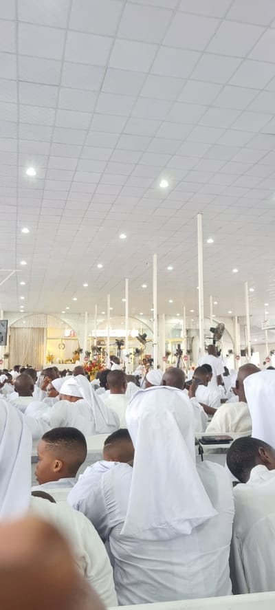 A photograph taken indoors shows a gathering of people in Anantigha, Nigeria. The room is characterized by white tiled walls and ceilings with evenly spaced light fixtures. The people are predominantly wearing white garments and are seated. Several of them appear to be looking down. Decorations, including flowers, are visible at the front of the gathering. There are also fans and what appears to be a sound system setup towards the front of the room.