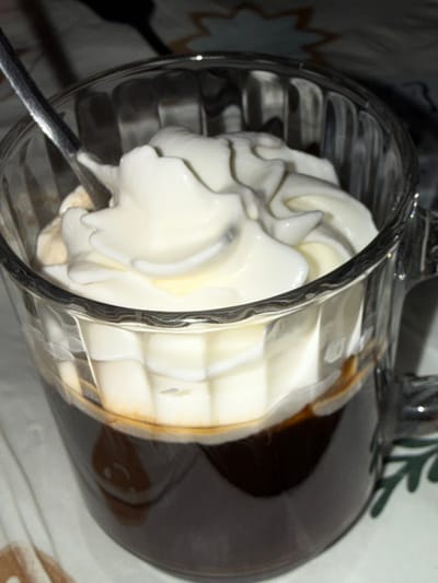 A close-up, dimly lit shot shows a glass mug filled with dark coffee topped with a generous swirl of whipped cream. A silver spoon rests inside the mug, partially submerged in the cream. The mug has a ribbed texture and sits on a patterned surface. A hint of green foliage is visible on the right side of the image. The location is not specified.