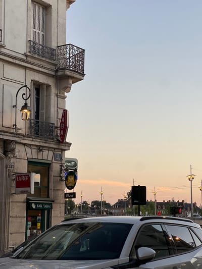 The image depicts an urban scene at dusk in Orléans, France, specifically at the corner of Rue Jean Mopeau and Pont George V.
**Scene Description:**
The sky exhibits a gradient from light blue overhead to a soft orange-pink at the horizon, indicating twilight. Numerous artificial lights are illuminated.
**Foreground:**
The front and passenger side of a light-colored (silver or white) sport utility vehicle (SUV) occupies the lower right portion of the frame, its windows reflecting ambient light and structural elements.
**Midground (Left):**
A multi-story building constructed from light-toned stone or stucco forms the corner of the intersection. The ground floor houses a "BAR TABAC," marked by a horizontal green and white sign. A prominent vertical red neon sign displaying "TABAC" is affixed to the building's corner. Below this, an ornate black wrought-iron street lamp with a lit, warm-toned bulb extends. Additional signage includes "PMU," a circular black and gold "Bar" sign with a stylized animal, and a partially visible "LA REP" sign (likely "La République du Centre"). A blue street sign, partially obscured, reads "RUE JEAN MOPEAU." The upper floors feature windows with closed white louvered shutters and decorative black wrought-iron balconies.
**Background (Right):**
The scene extends across a bridge, identifiable as Pont George V, which is lined with multiple illuminated, ornate streetlights. Indistinct human figures are visible on the bridge, appearing to walk. Distant buildings with dark, sloped roofs are visible on the far bank. A dark traffic signal pole with a red light is visible on the right side of the bridge.
No explicit interactions or specific actions are discernible among the limited human figures or within the vehicle.