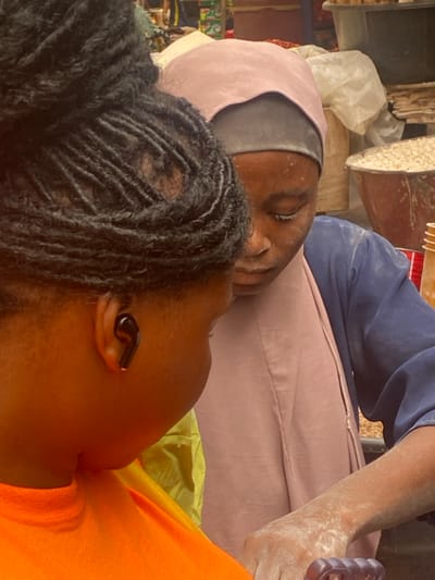 The image captures a close-up, rotated view of two individuals, likely resting or taking a break in what appears to be a work or market environment.
In the foreground, partially visible on the right, is a person with dark skin and black hair styled in short dreadlocks or braids. This individual is wearing a bright orange top and has a black wireless earbud visible in their right ear. Their face is seen in profile, looking towards the left side of the frame.
Behind and slightly to the left of the foreground person, another individual with a somewhat lighter skin tone is visible. This person is wearing a light pink head covering (possibly a hijab or shawl) over a dark grey or black undercap. Their eyes are closed, suggesting they are asleep or deeply resting. White powdery residue is noticeable on their face, particularly around the nose and forehead, and on their shoulders and arms. They are dressed in a blue long-sleeved garment over a light pink lower garment or fabric, both of which also show signs of the white powdery substance. Their right hand is visible, holding a purple object that resembles the back of a phone case or a small handled item, also covered in the white powder. The two individuals appear to be in close proximity, possibly leaning against each other.
The background is cluttered and suggests an indoor or partially covered outdoor setting, possibly a workshop, market stall, or storage area. Various objects are visible: white plastic bags, wooden planks or blocks, and other indistinct items. In the upper right background, there is a large, irregularly shaped, light-colored object, possibly a piece of equipment or structure, which also has some white residue on it. The general atmosphere, with the visible powder and functional background elements, hints at a place of activity or work where a powdery substance is handled, such as a flour mill, construction site, or a market dealing with certain dry goods. There is no visible text in the image. The lighting suggests daytime.