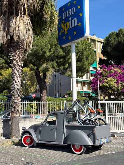 In Vallecrosia al Mare, Italy, a vintage grey Citroën 2CV pickup truck is parked on a cobblestone street. The truck has a customized rear bed fitted with a rack carrying two bicycles. The vehicles' wheels have white walls and red hubs. A tall palm tree with desiccated fronds stands to the left of the truck, casting a shadow. Behind the truck and fence, a building with green awnings is visible. A blue sign with yellow stars and the text "EURO Spin" is mounted on a pole to the right of the palm tree. The scene is under a clear blue sky on a sunny day.