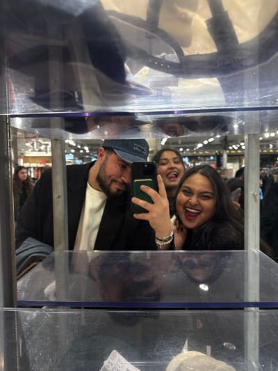 Three people pose for a selfie in a store. A man in a cap holds the phone, a woman beside him smiles widely, and another woman peeks from behind. The reflection in the glass reveals more of the store interior. Clothing, price tags, and other shoppers are visible. The photo is taken in a store fitting room.