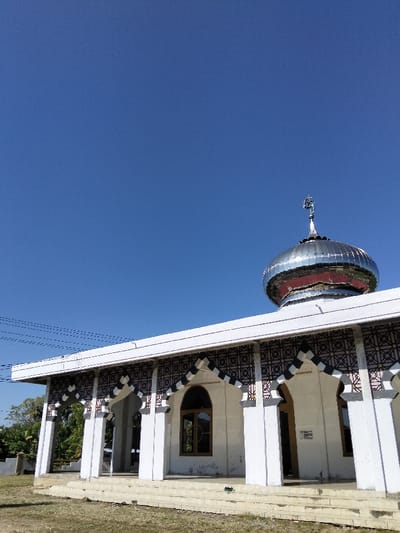 A mosque with a gleaming silver dome and a spire stands prominently against a cloudless blue sky. The building features a white facade with repeating arched entryways decorated with geometric patterns. A small rectangular window punctuates the wall. The mosque sits on a slightly elevated platform of stairs. The surrounding area appears to be a grassy field, suggesting a rural or suburban setting.