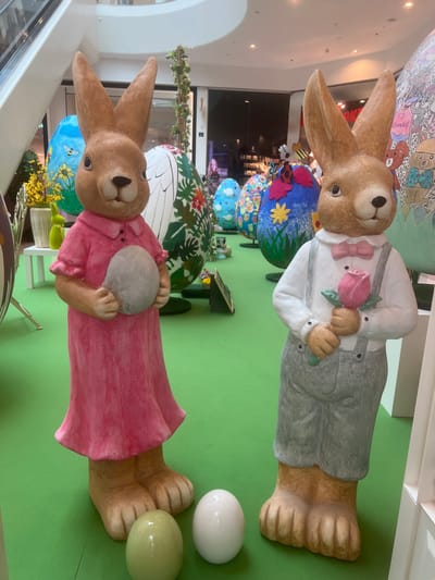 The image displays an indoor Easter holiday decoration exhibit located within a shopping mall in Vösendorf, Austria. Two large, anthropomorphic rabbit statues stand prominently on a bright green floor covering. The rabbit on the left, colored light brown, wears a pink collared dress and holds a smooth, pale grey egg. Adjacent to its base, a pale green egg and a white egg rest on the green surface. The rabbit on the right, also light brown, is dressed in a white collared shirt, a light pink bow tie, and grey overalls with white suspenders, holding a pink rose with green leaves in its hands. In the mid-ground, numerous oversized, ornately decorated Easter eggs are arranged, exhibiting diverse patterns including sunflowers with bees, tropical foliage, and abstract designs. The background features elements of the mall's architecture, including a partial view of an escalator in the upper left, high ceilings, and darkened storefronts with reflective surfaces, indicating a commercial retail environment.