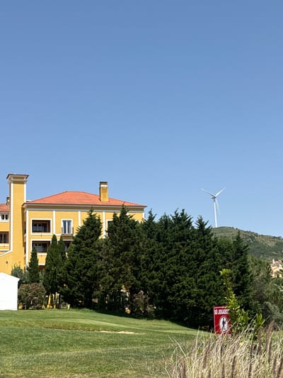 The image depicts a building with a terracotta-colored roof situated near a green lawn. A wind turbine is visible in the background. The sign "SD JOGADOR" is also visible. This is likely the location of Academia de Golf Runa, in Torres Vedras, Portugal.