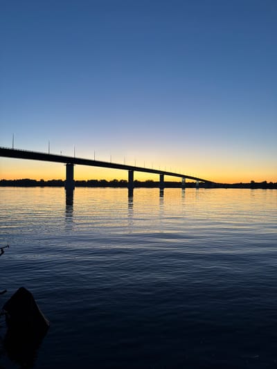 A wide shot captures the silhouette of a long bridge spanning a calm river at dusk, near Kambarka, Russia. The sky transitions from a clear, deep blue at the top to a warm, orange glow near the horizon, reflecting on the water's surface. The bridge, supported by multiple pilons, stretches across the frame, with streetlights visible along its length. A dark, silhouetted treeline marks the distant shore. In the foreground, the dark water is disturbed by gentle ripples, and a partially submerged black object is visible on the left side, likely part of a boat or floating debris. No people are present in the image.