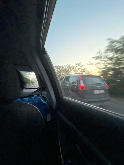 The image is taken from inside a moving vehicle, looking out the passenger side window. The foreground shows the interior of the car, including a dark-colored dashboard and a person's head and shoulder visible in the lower left corner, with a blue bag on their lap. The middle ground is dominated by a dark-colored SUV driving on the same road, with its brake lights illuminated. The SUV has a license plate visible, though the characters are blurry due to motion. Trees line the road on either side, appearing blurred due to the vehicle's movement. The background features a pale sky, transitioning from blue at the top to a yellowish hue near the horizon, suggesting either sunrise or sunset. The overall impression is a snapshot of a road trip on a clear day, possibly in or around Plodovitovo, Bulgaria, given the context.