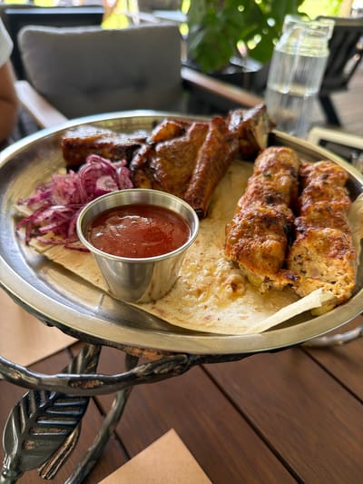 A metallic plate holding food is presented on a decorative stand with leafy designs. The plate contains two pieces of what appears to be grilled meat, possibly chicken skewers, and a portion of ribs. Alongside the meat, there is a small metal cup filled with a reddish sauce, a pile of thinly sliced red onions, and flatbread. The food is arranged on a large piece of flatbread. The background shows a blurred outdoor setting with furniture and greenery, suggesting a restaurant or cafe environment. The table surface is made of dark wooden planks.