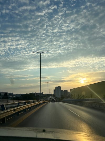 This media file captures a scenic view from a bridge, likely in Burtaiši, Montenegro. The orientation of the image suggests it was taken from a moving vehicle, possibly a car, looking towards the right. The primary subjects include a roadway with a single white line marking the lane, stretching into the distance. On the right side of the road is a yellow guardrail, and beyond that, a dark, possibly metal, railing.

The setting is a bridge overlooking a cityscape. Tall buildings, characteristic of an urban environment, are visible on the left side of the road, nestled against a backdrop of trees. The sky dominates the left half of the image, filled with a dynamic display of clouds. These clouds are small and scattered, creating a textured, almost rippled effect across the sky.

The time of day appears to be either sunrise or sunset, indicated by the warm, golden light that permeates the scene. The sun is partially obscured by clouds, casting a bright, diffused glow. The weather seems clear, with no rain or strong winds apparent.

A few cars are visible on the road, suggesting traffic is present but not congested. Two streetlights stand prominently on the left side of the roadway, their dark silhouettes contrasting with the bright sky.

There is no visible text in the image. The overall mood conveyed is one of peaceful travel and appreciation of a beautiful sky and cityscape. The angle and perspective imply the viewer is a passenger, observing the world outside the vehicle.
