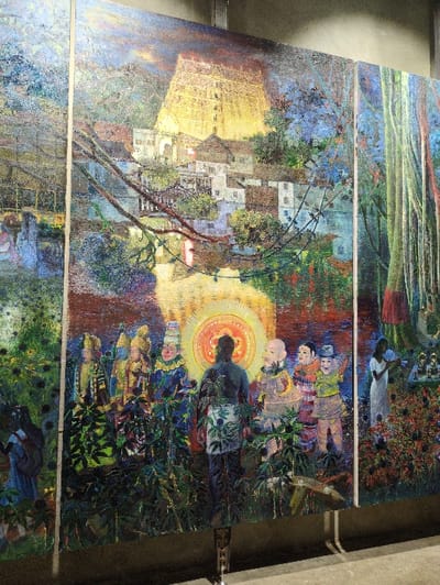 The image displays a large, multi-panel painting, likely a triptych, presented vertically on exhibition stands within an indoor space. The artwork employs a highly textured, vibrant style, utilizing rich blues, greens, yellows, reds, and browns.
The upper portion of the painting features a towering, golden-hued gopuram, characteristic of South Indian temple architecture, specifically resembling the Sree Padmanabhaswamy Temple. Below and around this structure, numerous white-walled houses with reddish roofs are nestled amidst dense green and blue foliage. A body of water, reflecting the sky and buildings, occupies the mid-ground, with dark branches extending over it. The sky above the temple appears dark and dramatic.
The lower and central sections of the painting depict a gathering of figures. A central figure, seen from behind, wears dark clothing with a light, leafy-patterned waist wrap, facing a prominent, radiant circular emblem featuring orange and red patterns. To the left of this central figure, several individuals with distinct headdresses and colorful, traditional garments stand amidst lush, dark green plants. To the right, other figures include a bald, bearded individual and another wearing a conical hat, also in colorful attire. The immediate foreground is dense with broad-leaved plants, partially obscuring the figures' lower bodies.
The rightmost panel features tall, slender trees with hanging roots, resembling banyan trees, extending vertically. In the lower right, a female figure dressed in white is positioned amidst red and green flora, appearing to interact with small objects on a low platform or altar.
The artwork is physically displayed in an indoor setting, with metal stands visible supporting the panels at the bottom. The location of the display is an indoor gallery or exhibition space, such as those found in the city of Kochi, India.