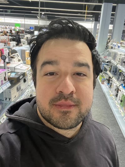 The image captures a selfie of an adult male, likely taken indoors in a retail store environment. The man's face is prominent, filling the upper-right portion of the frame (when oriented correctly). He has dark, wavy hair, and a short beard or heavy stubble covering his chin and upper lip. His eyes are dark, and he has a neutral expression, looking directly at the camera. He is wearing a dark, possibly black or charcoal gray, hooded sweatshirt or jacket, with the hood visible around his neck and shoulders.

The background reveals the setting to be a store with numerous shelves stocked with various packaged goods. On the left side of the frame, shelves extend into the distance, displaying items that appear to be electronics or household appliances in their boxes, some with visible labels and price tags. The shelves are well-lit by overhead artificial lighting, likely fluorescent, which casts reflections on the shiny surfaces of some products and the store floor. A green sign with white text is partially visible on one of the displays in the background, though the text itself is unreadable. On the right side of the frame, more shelves packed with similar products are visible, receding into the background. The overall impression is that of a modern department store or an electronics retail outlet. The image is rotated 90 degrees clockwise from its likely original orientation.