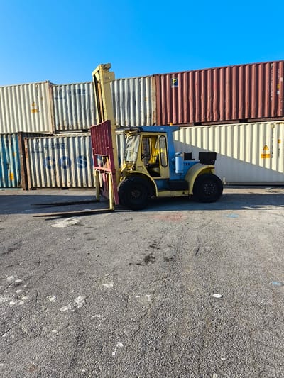 A yellow and blue forklift is parked on a paved surface in front of stacked shipping containers under a clear blue sky. The forklift has a red fork attachment raised. The shipping containers are stacked in rows, with some white, blue, and red containers visible. The ground appears to be asphalt or concrete and shows some signs of wear and tear, with light-colored splatters and debris. The scene suggests an industrial or port environment. The location in the Municipal Unit of Inofyta, Greece, places this scene within an industrial zone known for manufacturing and logistics operations.