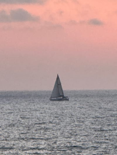 A sailboat with a triangular sail is visible on water. The sky to the left appears tinted with pink or orange hues, possibly during sunrise or sunset. The water's surface exhibits ripples, suggesting a gentle breeze. The overall image has a serene and calm atmosphere, with the sailboat as the central subject.