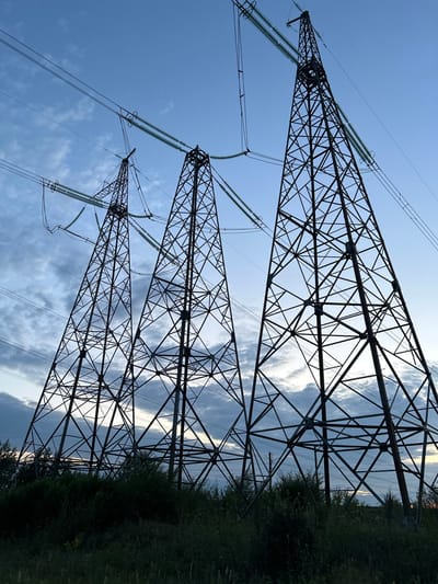 Three towering, dark metal lattice electricity pylons dominate the frame, stretching upwards against a twilight sky with scattered clouds. The pylons are silhouetted, their intricate geometric structures clearly visible. Power lines, some taut and others slightly sagging, are strung between them and disappear out of frame. The pylons are set in a landscape of dense, dark green foliage and grass in the foreground, with hints of distant trees and a faint glow on the horizon. The scene is set at dusk or dawn, with the sky transitioning between blue and darker hues. The location is specified as the city of Belozerki, Russia.