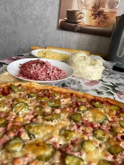 The image displays a close-up of a freshly baked pizza with toppings of pickles, ham, and melted cheese. In the background, there are three white plates, one containing shredded cheese, another with chopped ham and sausage, and a third with a ball of dough wrapped in plastic. A wooden rolling pin rests horizontally on top of the plate of shredded cheese. To the right, a black electric kettle with a visible water level indicator is present. Above the kettle, a framed picture depicts a teapot and a teacup. The scene is set on a table covered with a floral-patterned tablecloth. The background wall has a textured gray finish. The overall setting appears to be a kitchen or dining area. The image does not provide any information about the city of Oktyabrsky, Russia.