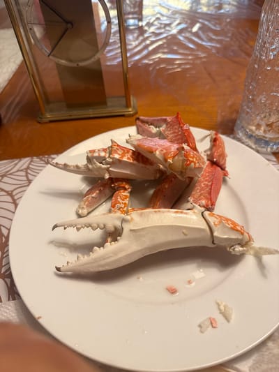 A close-up, overhead shot shows a white plate filled with cooked crab legs, some pieces cracked open, on a wooden table. A vintage-style brass and glass clock is visible in the background, to the left of the plate. To the right, a part of a textured glass bottle or vase can be seen. The scene is lit by artificial light, casting shadows and reflections. The overall setting suggests a meal, possibly in a domestic environment. There is no information in the image to suggest it is located in Bonn, Germany.