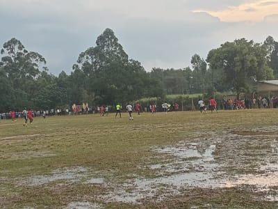A soccer match unfolds on a waterlogged field beneath an overcast sky. Players in red and white jerseys are spread across the muddy pitch. Spectators line the sidelines. Trees form a backdrop to the game. A building is visible on the right. Puddles reflect the sky and nearby trees.