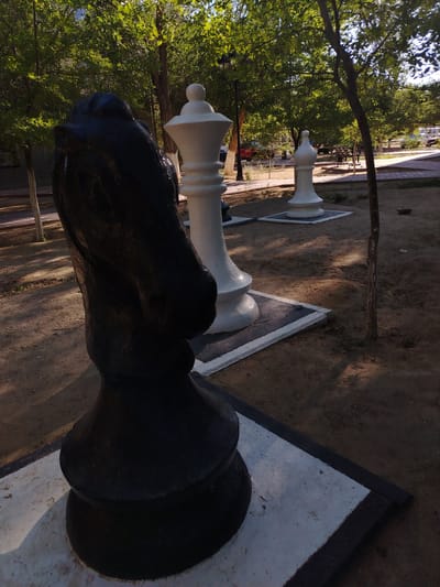 This image captures an outdoor scene featuring oversized chess pieces, likely a public art installation or a park feature. In the foreground, a large, black chess knight, sculpted to resemble a horse's head, stands prominently. Behind it, two white chess pieces, appearing to be pawns or perhaps bishops, are visible. The chess pieces are positioned on square bases with white and dark borders.
The setting is a park-like area with sandy ground, scattered small stones, and sparse vegetation. Several trees with lush green foliage are scattered throughout the scene, casting dappled shadows on the ground, suggesting it is daytime, likely late afternoon or early evening due to the warm, golden light. In the background, a paved walkway and benches can be seen, hinting at a recreational area. Further in the distance, there are hints of buildings and parked vehicles, indicating an urban or semi-urban environment.
The overall mood is serene and peaceful, with the sunlight filtering through the trees creating a tranquil atmosphere. No people are visible in the frame. There is no visible text in the image. The location context provided is Baikonur, Kazakhstan.