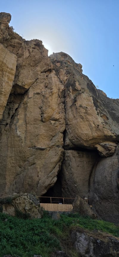 This is a vertical, eye-level, full shot photograph capturing a section of the cliff face and the entrance to a cave at the Areni-1 cave complex, located in the Vayots Dzor Province of Armenia. The sun is high in the sky, partially obscured by the upper cliff, casting a bright light and creating some blown-out areas in the sky. The rock formations are rugged and textured, appearing to be composed of sedimentary rock with layers and fissures.

At the bottom of the frame, a wooden platform with a simple railing has been constructed to provide access to the cave entrance. The cave opening is a dark, irregular shape set into the base of the massive rock face. Lush green vegetation covers the ground in the foreground, leading up to the platform and the base of the cliffs.

In the far upper right of the image, a small bird is visible in flight against the bright blue sky. There are no people visible in the scene. The overall impression is one of a natural geological formation adapted for human access and exploration.