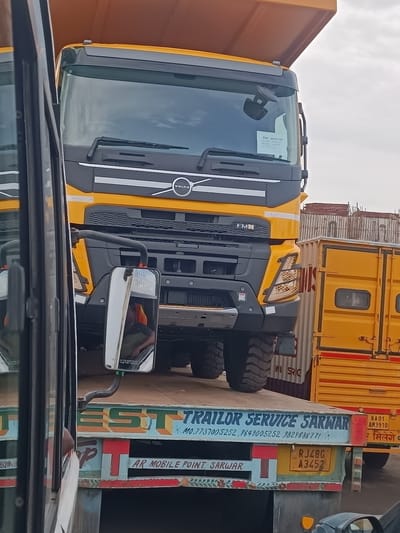 In Balanagar, India, a yellow Volvo truck is parked, its large orange cargo bed tilted upwards. The truck's front is visible, with its distinct Volvo logo and "FM" badging. On the windshield, a white sticker with text is affixed. The truck is positioned next to a yellow shipping container. In the foreground, a portion of another vehicle is visible, reflecting the scene. Below the Volvo truck, the rear of a flatbed truck is visible, adorned with colorful painted text. The text on the flatbed reads "TRAILOR SERVICE SARWAR" and also displays a phone number: "MO.7737005252, 9849005252, 9829886771". Another line of text below reads "AR MOBILE POINT SARWAR". A license plate with "RJ48G A3452" is also visible on the flatbed. The environment appears to be an outdoor industrial or logistical area, with construction materials visible in the background. The sky is overcast.