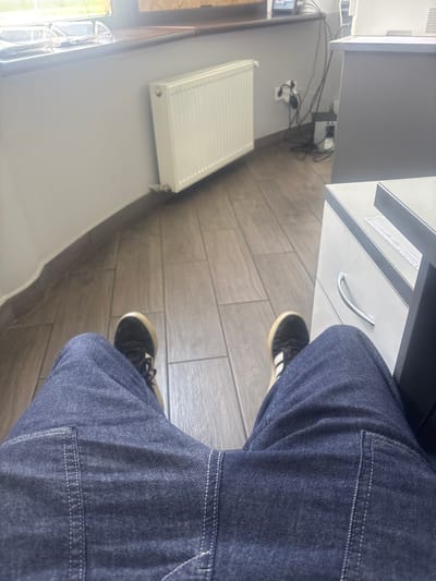 This media file is a first-person perspective video showing a person sitting at a desk. The subject is wearing dark blue denim jeans and black and white sneakers. The scene is an indoor setting, likely an office or study, as indicated by the desk, filing cabinet, and what appears to be a radiator. The flooring is made of wooden planks.
Notable details include the presence of a white radiator mounted on a light grey wall, and a grey filing cabinet with a silver handle. Cables and a power strip are visible near the base of the filing cabinet. A window is partially visible on the left side of the frame, suggesting daylight, but the time of day and weather are not definitively discernible. There is no discernible text within the video. The activity is static, suggesting the person is seated and possibly working or resting. The mood is neutral. The location is specified as Pereiaslavske, Ukraine.