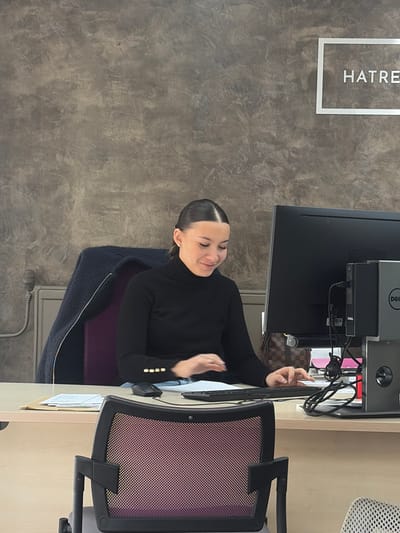 The scene is an interior office located in Cagnes-sur-Mer, France. A woman with dark hair pulled back, wearing a black turtleneck shirt with gold buttons on the cuffs and light-colored trousers, is seated at a light-colored desk. She is actively typing on a black keyboard, positioned in front of a black Dell computer monitor. Her gaze is directed downwards towards the keyboard/documents, and she exhibits a slight smile. A black computer mouse rests on the desk to her left. Various documents are present on the desk, with some contained within a light-brown folder. A dark blue coat with a yellow trim is draped over the back of a purple upholstered office chair situated to the left of the woman. A brown Louis Vuitton patterned bag is partially visible behind the monitor. The background wall features a dark gray, textured finish. A silver-framed sign bearing the partial text "HATRE" is mounted on the wall in the upper right. A light-colored pipe or radiator fixture is visible on the wall to the far left. The back of a dark-framed office chair with a purple mesh insert is present in the foreground.