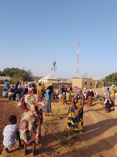 This image depicts an outdoor gathering in Bahit, Nigeria, under a clear blue sky. A diverse group of approximately 50-60 individuals, predominantly women and some children, are present on dry, reddish-brown dirt ground. Many are seated or kneeling, while others stand, largely facing towards the right side of the frame or into the distance. Individuals wear brightly colored, patterned traditional dresses, and many women have head coverings.
In the foreground, a young boy kneels to the left, and a woman in a brown, green, and pink patterned dress stands with her back to the camera. Another woman, similarly facing away, wears a blue and yellow patterned dress with a light-colored cloth draped over her head.
The background features several structures: a building with a light-colored pyramidal roof, an incomplete brick structure, and other distant buildings. A prominent red and white lattice communication tower with antennas stands tall against the sky. Sparse trees and dry vegetation are scattered throughout the scene. The overall environment appears dry and sunny, consistent with a rural or semi-urban setting.