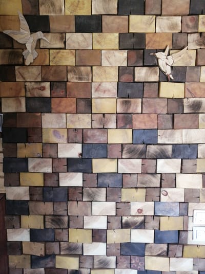 The image displays a close-up, eye-level view of a wall art installation composed of numerous wooden blocks of varying colors and wood grain patterns. The blocks are arranged in a brick-like pattern, creating a textured and rustic surface. Two decorative wooden bird cutouts are affixed to the wall. The left bird cutout, larger and positioned towards the top left, is light in color with visible wood grain. The second bird cutout, smaller and located in the upper right quadrant of the image, is also light-colored and appears to be made of layered wood pieces. The background consists of the interlocking wooden blocks, some exhibiting dark, weathered finishes, while others show lighter natural wood tones, including rings and knots. The lighting suggests an indoor setting with diffuse illumination, highlighting the three-dimensional quality of the wood. The image is focused on the wall art itself, with no discernable environment beyond the artwork. The location in Yemelyanovo, Russia is not visually represented within the provided image.