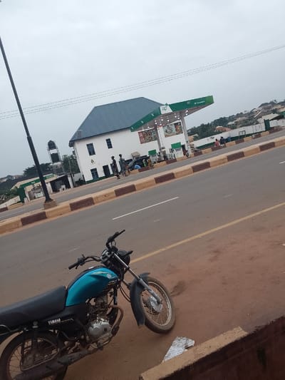 The image depicts an outdoor scene in Ijara, Nigeria, featuring a paved road alongside an "ap" branded gas station under an overcast sky. In the immediate foreground, a teal and black Yamaha motorcycle is parked on a reddish-brown dirt shoulder. The motorcycle is positioned facing left, with its front wheel slightly turned. To its right, a low concrete barrier is partially visible.
The midground is dominated by a two-lane asphalt road with a yellow centerline and white edge lines. Brown and beige concrete road barriers separate the road from the gas station on the right. The gas station includes a white, two-story building with a distinctive blue corrugated metal roof and an "ap" branded green and white canopy structure covering multiple fuel pumps. Approximately 10-15 individuals are observed in and around the gas station area; some appear to be walking near the building and pumps, while others are less clearly defined. A tall, black utility or street light pole stands on the left side of the road, and behind it, a black elevated water tank is visible.
In the background, rolling hills covered with sparse vegetation and residential structures extend under the uniformly gray, cloudy sky. Overhead, multiple utility lines traverse the frame.