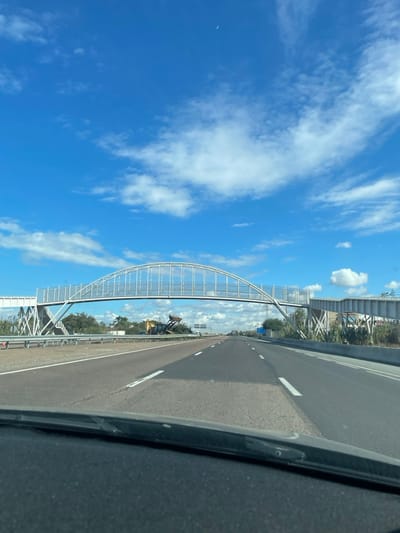 The image depicts a multi-lane highway in Skhirat, Morocco, viewed from the interior of a vehicle looking forward. A prominent white metal pedestrian overpass with an arched truss structure spans across the highway. The overpass features transparent or mesh panels along its sides and stair access on both the left and right sides of the road.
To the left of the highway's central median, beneath the overpass, a yellow articulated front-end loader or excavator is visible amidst a pile of debris or cleared vegetation. The loader's bucket is elevated. Orange traffic cones and white barriers are positioned on the median near the construction equipment.
The highway itself is asphalt, with white dashed lane markings. The pavement shows variations in tone, indicating potential repair or different material sections. The sky is bright blue with scattered white cumulus and cirrus clouds. A faint crescent moon is visible in the upper center-right portion of the sky. Sparse green vegetation, possibly shrubs or small trees, lines the shoulders of the highway. No people are visible within the frame.