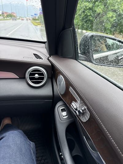 The image is an interior shot of a car driving on a wet road in Oktyabrskoye, Russia. The perspective is from the passenger seat, looking forward and to the right. Raindrops are visible on the windshield and side window, indicating inclement weather. The car's interior features dark brown leather upholstery with stitching, wood trim accents, and chrome detailing on the door controls. A person's legs, wearing blue jeans and a light-colored shoe, are visible in the lower left of the frame, resting on the car's floor mat. Outside the car, the road is visible, with other vehicles in motion and buildings lining the street. Trees and vegetation are also seen along the roadside. The sky appears overcast.